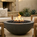 A round concrete fire pit bowl with black fire rocks and active orange flames, situated outdoors between patio chairs in a backyard setting.