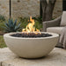 A round concrete outdoor fire pit bowl filled with black lava rocks and active orange flames, set on a stone patio with a wicker chair and garden plants in the background.