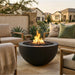 A round black fire pit table with an active flame, set on a stone patio between outdoor seating in a residential backyard setting.