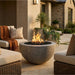 Outdoor patio with round concrete fire pit, chairs, and decorative plants.