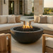 A round dark gray outdoor fire pit bowl filled with black stones and glowing orange flames, placed between cream outdoor sofas on a stone patio in front of a modern house.