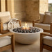 A round concrete fire pit with gray stones and a visible flame, placed between beige patio armchairs in a stone patio setting.