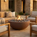 A round outdoor fire pit with a terracotta-colored bowl and glowing flames, filled with dark river rocks, set on a stone patio between modern wooden patio chairs and a sofa in a warm residential backyard setting.