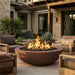 A round dark brown fire pit with black stones and bright orange flames, situated on a stone patio in a modern outdoor living room setting with wooden chairs and plants.