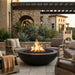 A round black fire pit with a glowing fire, set in a modern outdoor patio with beige lounge chairs, stone walls, and plants.
