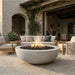 A round concrete fire pit with black fire rocks and a visible flame, placed on a stone patio in a modern outdoor living room setting with upholstered seating and a stucco building.
