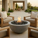 A round concrete fire pit table with a glowing flame and dark river rocks, set on a tiled outdoor patio between two wooden armchairs with beige cushions.