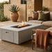 A rectangular concrete fire pit table with a visible flame and dark decorative stones inside, placed on a stone patio beside a beige sofa with pillows and a draped throw blanket.