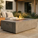A rectangular concrete fire pit table with a live flame is situated on a stone patio in front of a modern outdoor sofa and landscaping.
