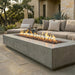 A rectangular concrete outdoor fire pit table with active flames, positioned in front of a beige patio sofa and lounge chair on a stone patio in front of a house.