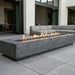 A modern rectangular concrete fire pit with active flames, set on a gray stone patio in front of a contemporary outdoor sectional sofa.