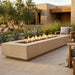 A long rectangular outdoor fire pit with visible flames, set on a patio in front of a modern house with plants and seating visible.
