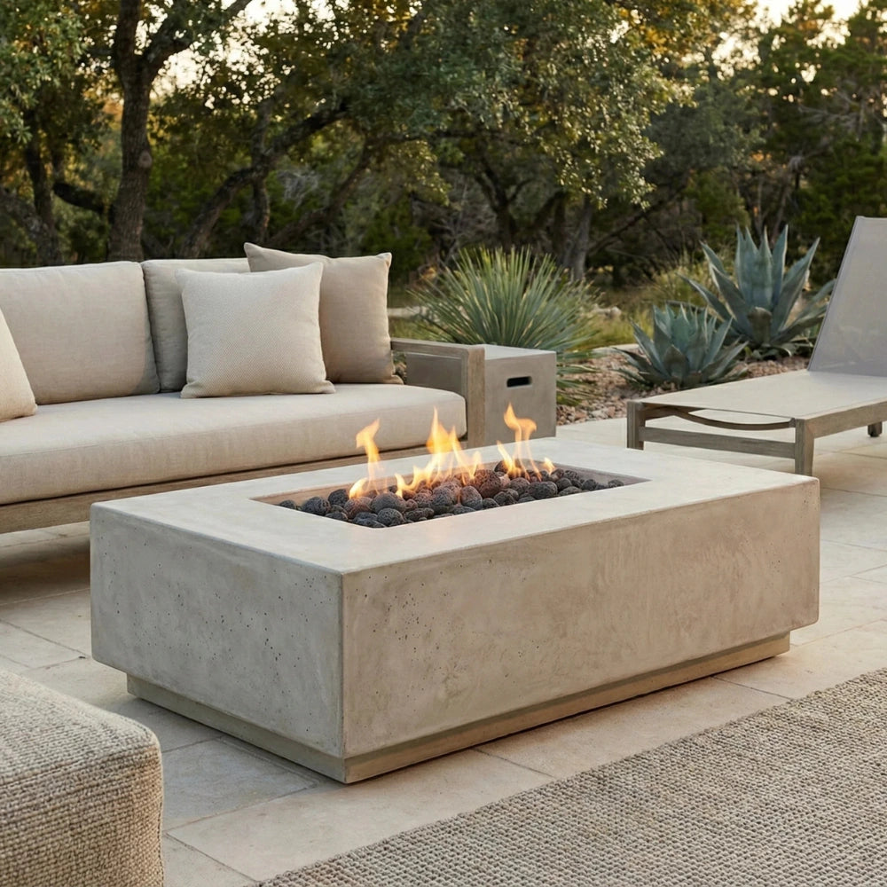 A rectangular concrete fire pit table with a live flame and dark river rocks, set on a stone patio in an outdoor patio setting with a beige sofa and lounge chair.