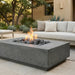 A square concrete fire pit table with a lit flame and dark river rocks, set on a patio in front of a light beige outdoor sofa and house exterior.