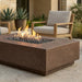 A rectangular concrete fire pit table with a square fire opening filled with dark river rocks and active orange flames, set on a stone patio with outdoor lounge chairs and potted plants in the background.