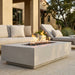 A rectangular concrete outdoor fire pit table with a lit flame and dark river rocks, set on a stone patio in front of a modern outdoor lounge sofa.