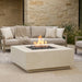Outdoor patio setting with a square concrete white fire pit table and sofa against a stone wall.