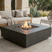 A square concrete fire pit table with a lit flame and dark stones, set on a patio in front of a beige outdoor sofa.