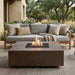 A rectangular concrete fire pit table with a lit fire is shown in front of a modern outdoor sofa on a patio. The scene includes a house exterior, plants, and trees in the background.