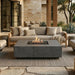 A modern outdoor patio scene with a rectangular concrete fire pit table in the foreground, a light beige sectional sofa with matching pillows, a woven rug, stone flooring, and a stone wall background.