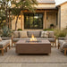 Outdoor patio conversation set featuring a rectangular concrete fire pit with a visible flame, paired with a matching three-seat sofa and armchairs on a woven rug in front of a stone house.