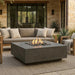 Outdoor patio with grey square concrete fire pit table, sofa, and chairs in a stone-walled setting