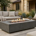 A rectangular concrete fire pit table with a fire burning inside, set in an outdoor patio area next to a beige sectional sofa.