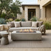 Outdoor patio furniture set featuring a light beige sectional sofa with matching throw pillows, a rectangular concrete fire pit table with active flames, a matching concrete side table, and a woven area rug in a modern backyard setting.