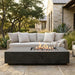 Outdoor patio furniture set featuring a light beige sectional sofa with matching pillows and a dark charcoal rectangular fire pit table with active flames, set on a stone patio in front of a stone house with trees in the background.