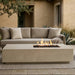 A rectangular concrete fire pit table with a live flame is placed in front of a matching beige outdoor sofa on a patio in front of a stucco house.