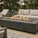 A rectangular outdoor fire pit table with a dark gray concrete-look finish, filled with small gray stones and active orange flames, set in a patio lounge area with a light beige sofa and outdoor furniture in the background.