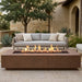 A rectangular concrete fire pit table with a visible flame sits outdoors in front of a light gray outdoor sofa on a stone patio, with a stucco house, tree, and plants in the background.