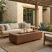 A rectangular outdoor fire pit table with a brown metal finish and visible flames, placed in front of a beige outdoor sofa on a woven rug in a patio setting.