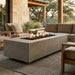 A rectangular concrete fire pit table with a live flame and dark river rocks, styled on a woven rug in a modern outdoor patio setting with a sectional sofa and wooden chairs.