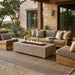 Outdoor patio furniture set including a rectangular concrete fire pit table, a three-seat sofa, a matching armchair, and a chaise lounge with beige cushions and light wood frames, arranged on a woven rug in front of a stone house.