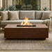 Outdoor rectangular concrete fire pit table with flames in front of a beige sofa and decorative pillows.