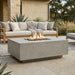 A square concrete fire pit table with a visible flame sits on a stone patio in front of a light wood outdoor sofa with beige cushions and a throw blanket.