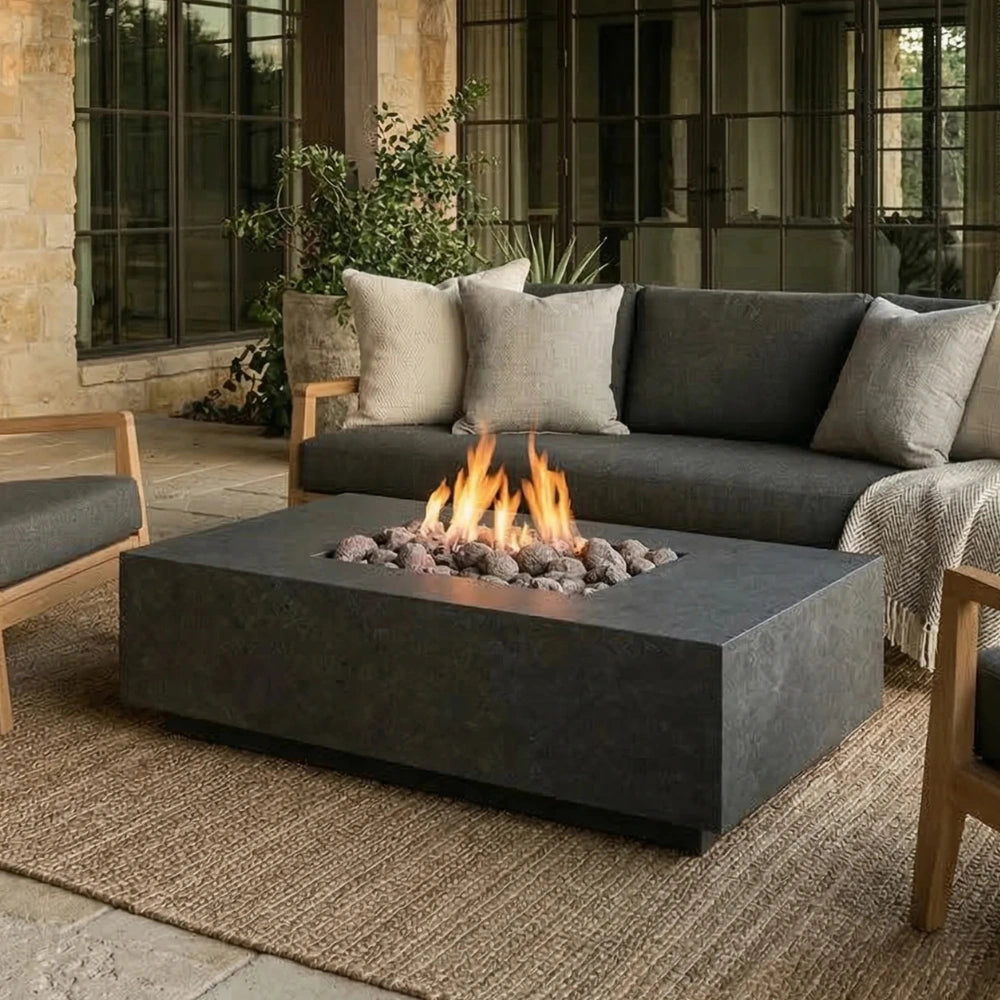 A modern rectangular outdoor fire pit table with a dark gray concrete finish, active flames, and decorative stones, positioned in front of a dark gray sectional sofa on a woven jute rug.