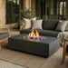 A modern rectangular outdoor fire pit table with a dark gray concrete finish, active flames, and decorative stones, positioned in front of a dark gray sectional sofa on a woven jute rug.