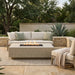 Outdoor patio furniture set featuring a beige sectional sofa with light green pillows, a matching rectangular fire pit table with visible flames, and a beige armchair with a draped throw blanket, arranged on a woven rug on a stone patio with trees and plants in the background.