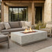 Outdoor patio furniture set featuring a rectangular concrete fire pit table with active flames, a gray sectional sofa with neutral throw pillows, and a matching armchair, arranged on a woven rug in front of a stone house.