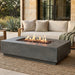 A rectangular concrete fire pit table with a live fire burning inside, set on a woven outdoor rug in front of a beige patio sofa on a stone patio.