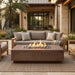 Outdoor patio furniture set featuring a rectangular concrete fire pit table with active flames, a three-seat sectional sofa with taupe cushions, and matching armchairs in a modern stone house setting.