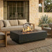 A modern rectangular outdoor fire pit table with a black concrete-look body and active flames, placed on a woven rug in front of a beige outdoor sofa on a stone patio.
