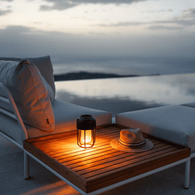 A solar-powered black portable outdoor lamp placed on a square wooden outdoor coffee table beside white sectional sofa