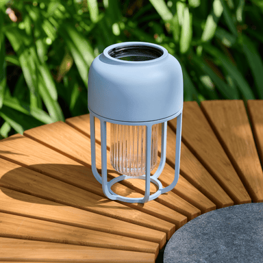 A solar-powered white portable outdoor lamp placed on a round wooden outdoor dining table beside plants