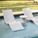Two light gray in-pool chaise loungers and a round side table on a pool sunshelf with a green lawn area in the background.