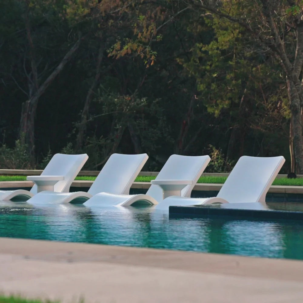 Four white in pool chaise loungers and two side tables partially submerged in a pool, surrounded by trees.