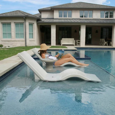 A woman in a wide-brimmed hat on a pool chaise lounger, reading a book while partially submerged in water, with a modern house nearby.