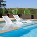 Two white tanning ledge chairs and a round side table partially submerged in a pool, with green plants in square planters behind.
